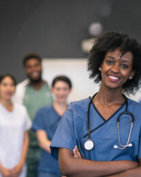 Portrait of a team of nurses and doctors. The multi-ethnic medical professionals are standing in a conference room in a hospital. The confident group is looking directly at the camera. They have their arms crossed and are smiling. A female doctor of African descent is standing at the front of the group.